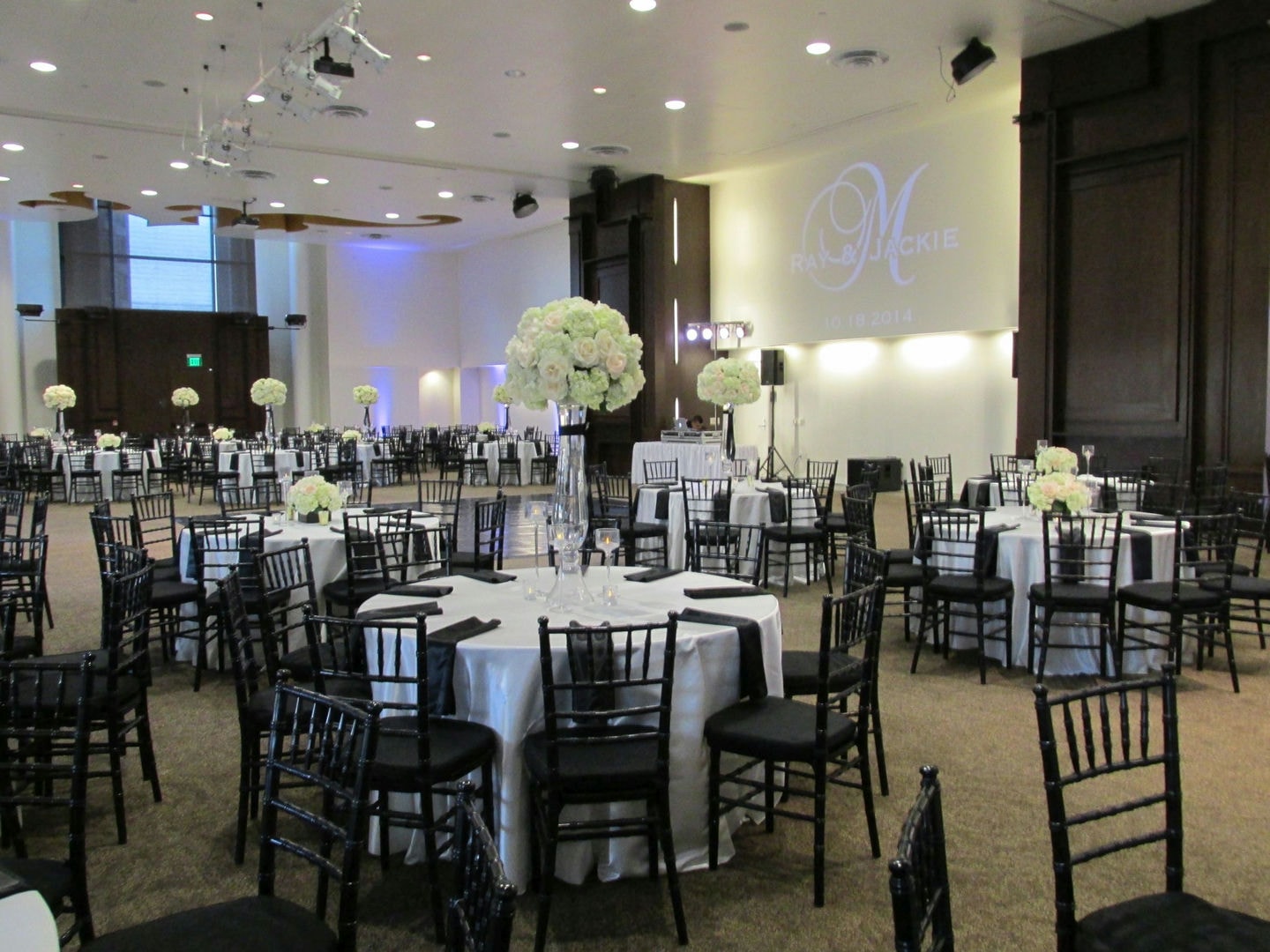 A large room with round tables, black chairs, and white tablecloths. Floral centerpieces are visible.