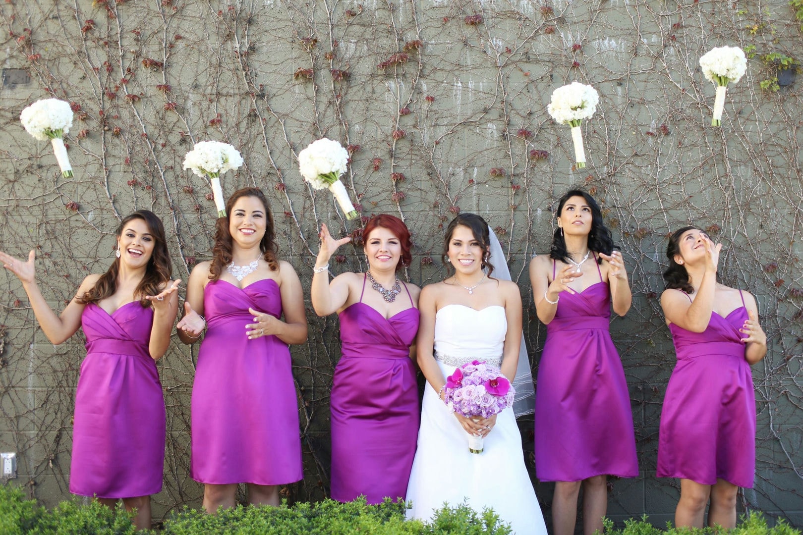 bridesmaids in purple dresses, a bride in white, and white bouquets thrown in the air.