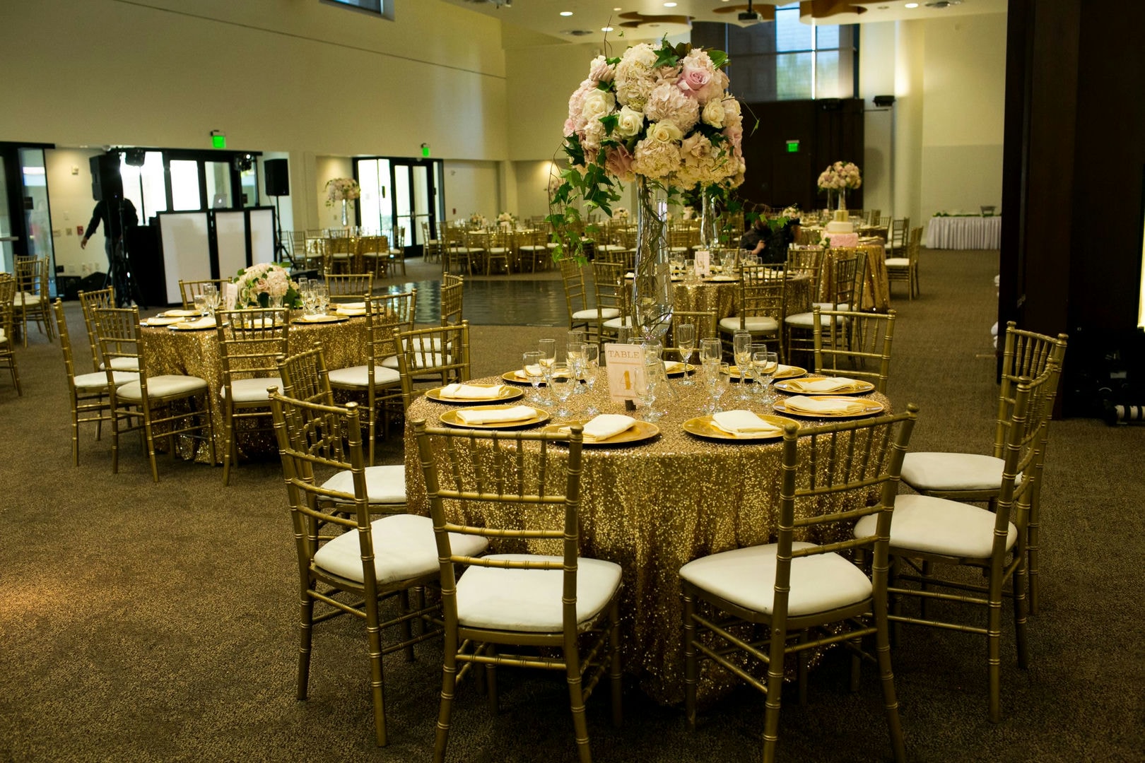 A banquet hall with gold tables and chairs, and large floral centerpieces.