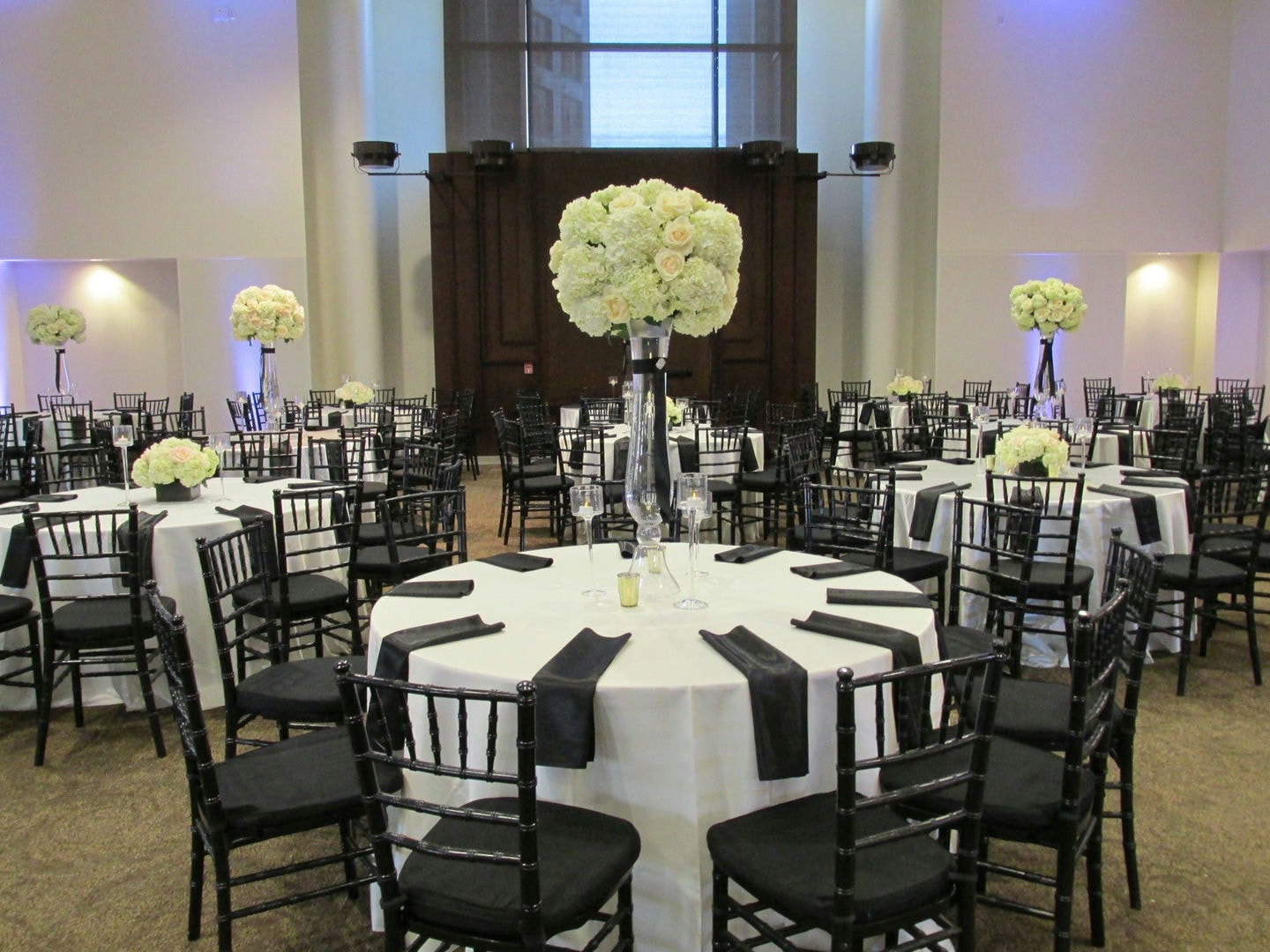 A large room with round tables, black chairs, and white tablecloths. Floral centerpieces.