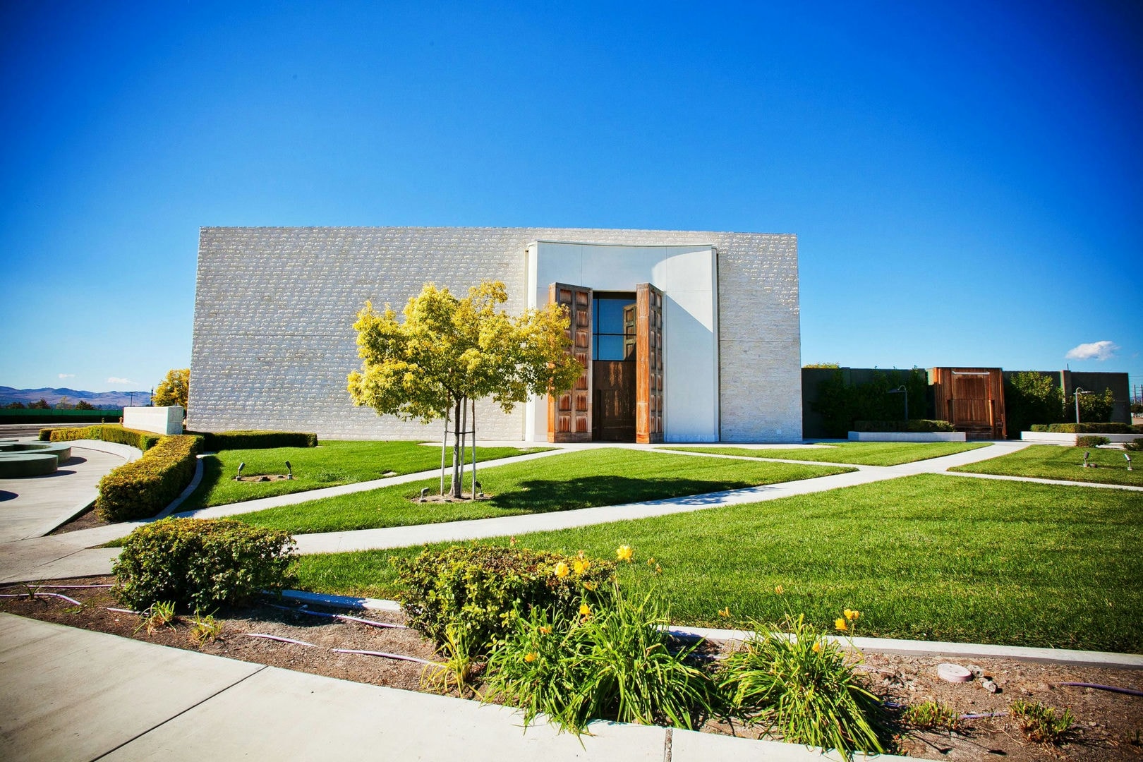 A light-colored building with a light-brown door, surrounded by green grass and landscaping.