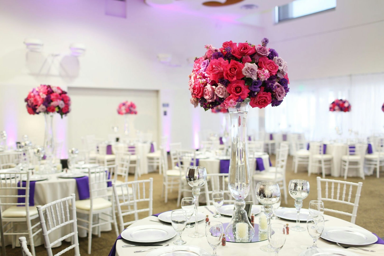 A banquet hall with white tables, chairs, and purple accents. Tall flower arrangements are on the tables.