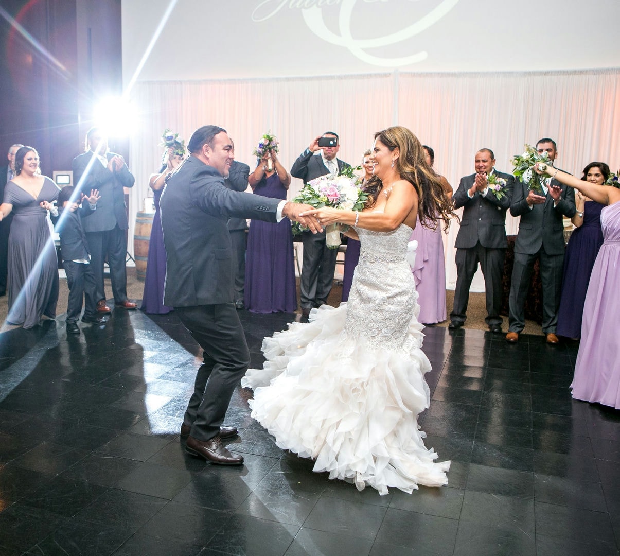 A bride and groom are dancing on a dark-tiled floor. Many people are watching.