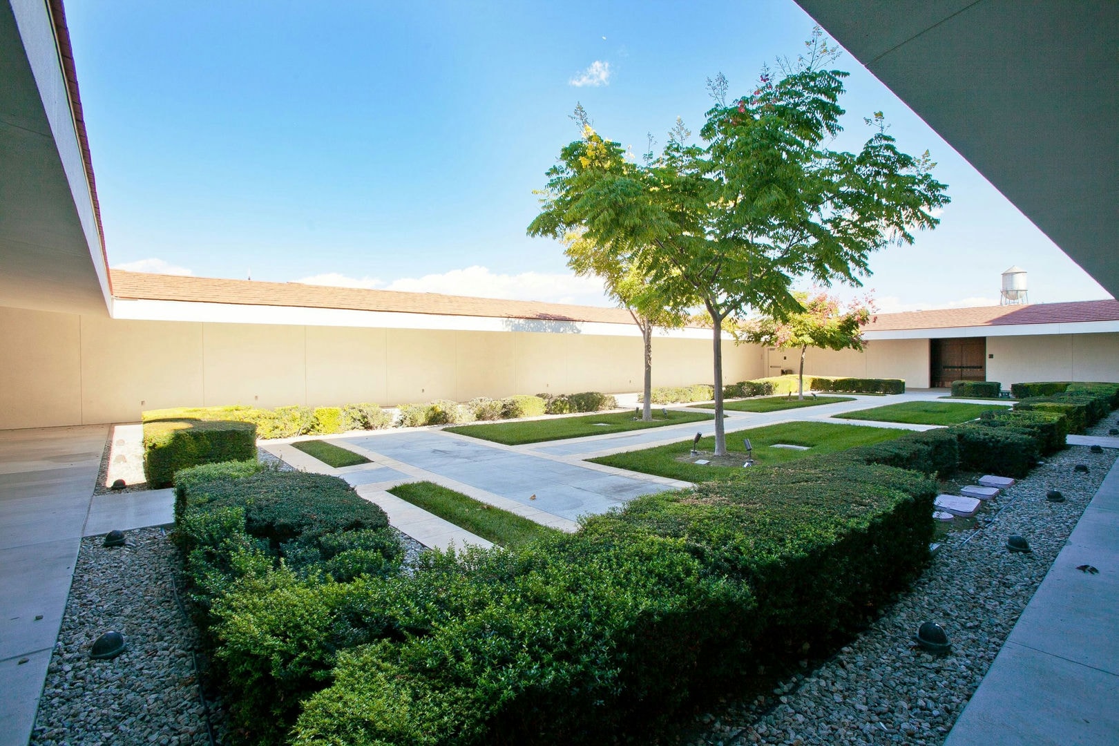 A courtyard with light beige walls, green hedges, trees, and light gray paved walkways.