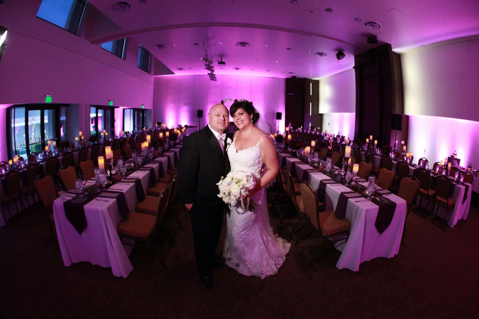 A wedding reception hall with tables, chairs, and a bride and groom. Purple lighting.