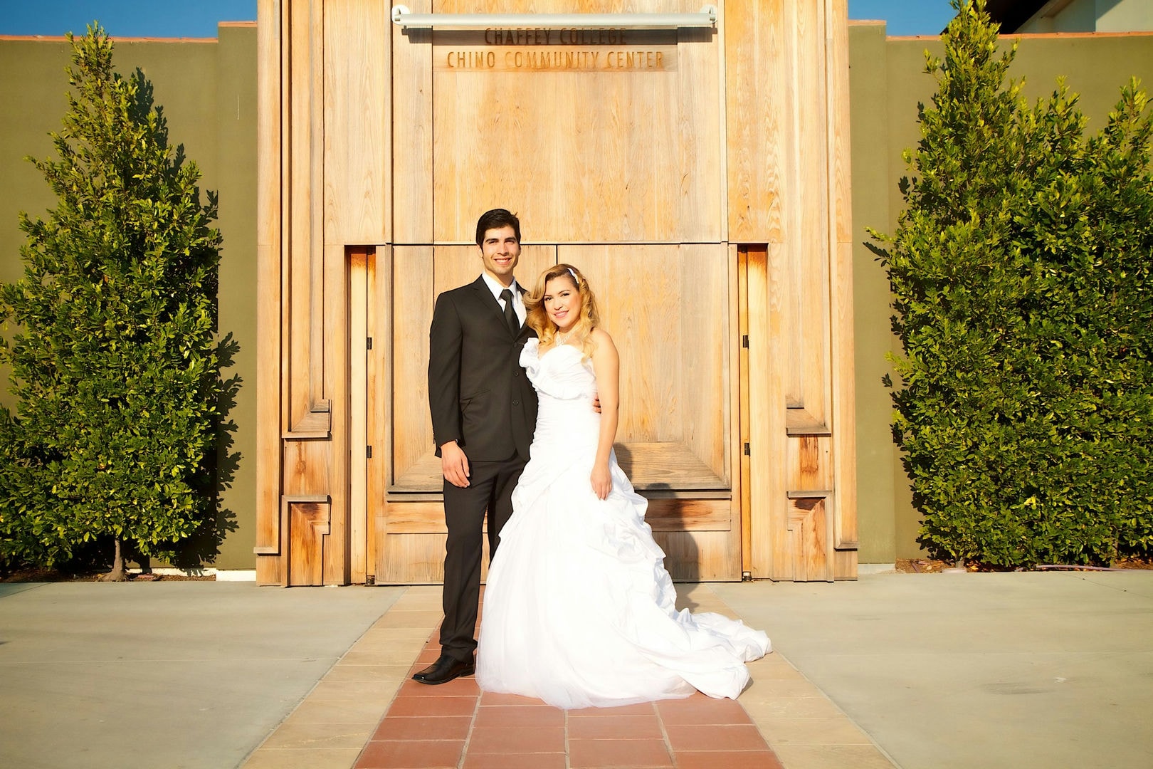 A couple in wedding attire stands in front of a light brown wooden building. 