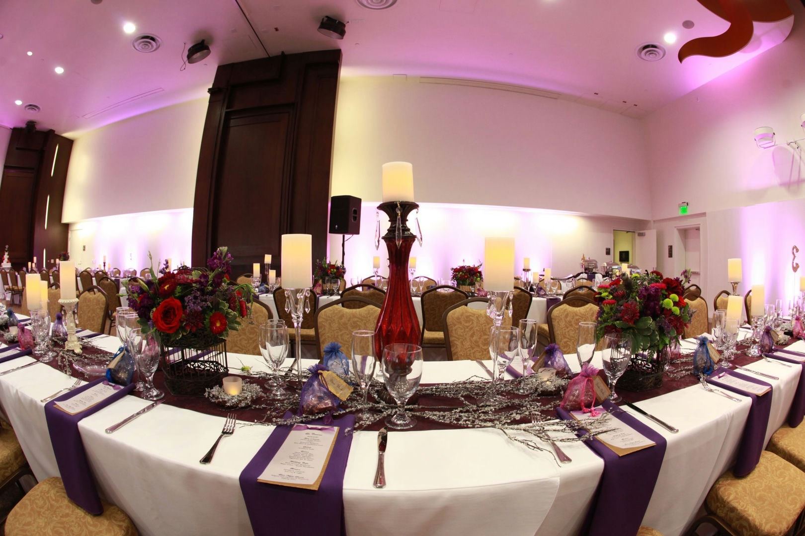 A banquet hall with long tables, white linens, purple accents, and floral centerpieces. Several people are seated.