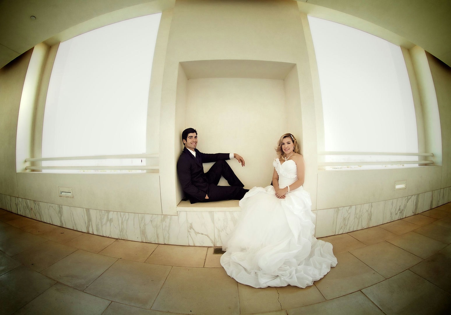 A bride and groom are sitting on a light beige stone ledge. They are wearing formal attire