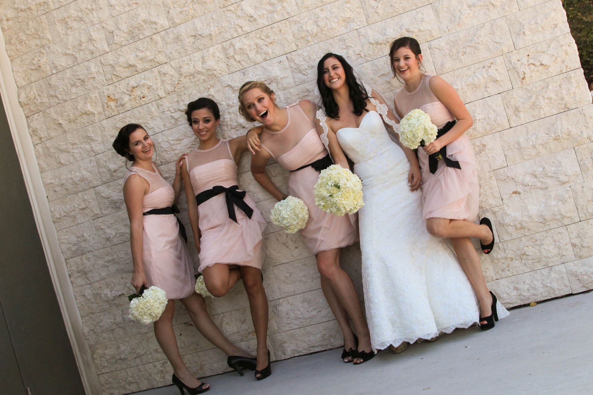 A bride and several bridesmaids in light pink dresses and a white wedding dress, posing in front of a light beige stone wall.