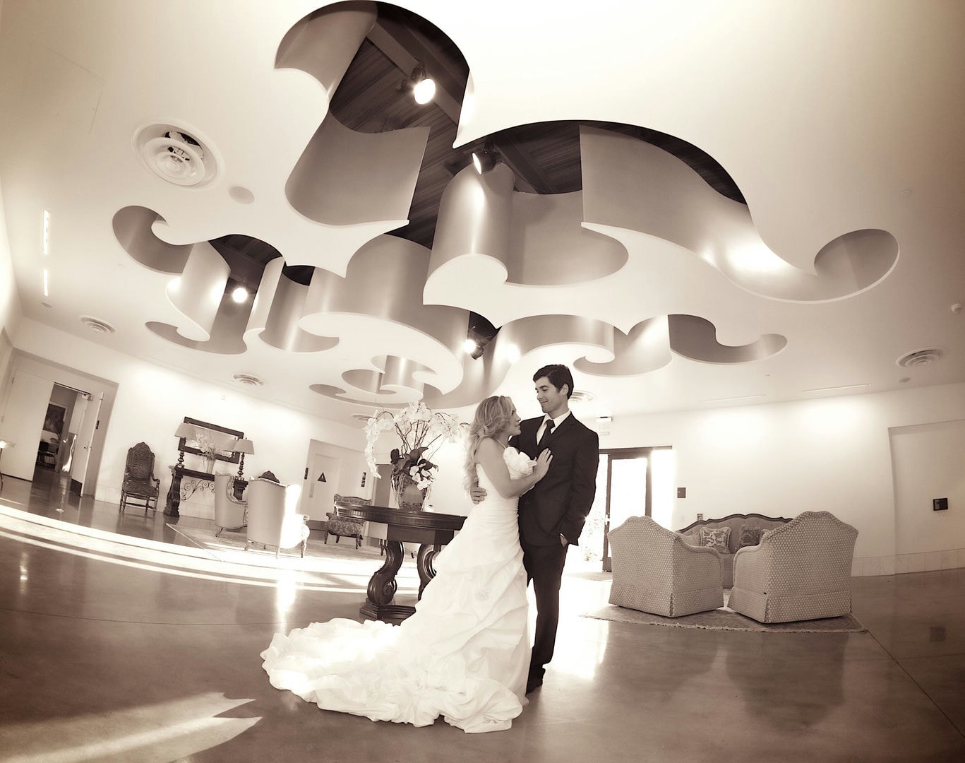 A wedding couple stands in a light-filled hall with a decorative ceiling and seating.