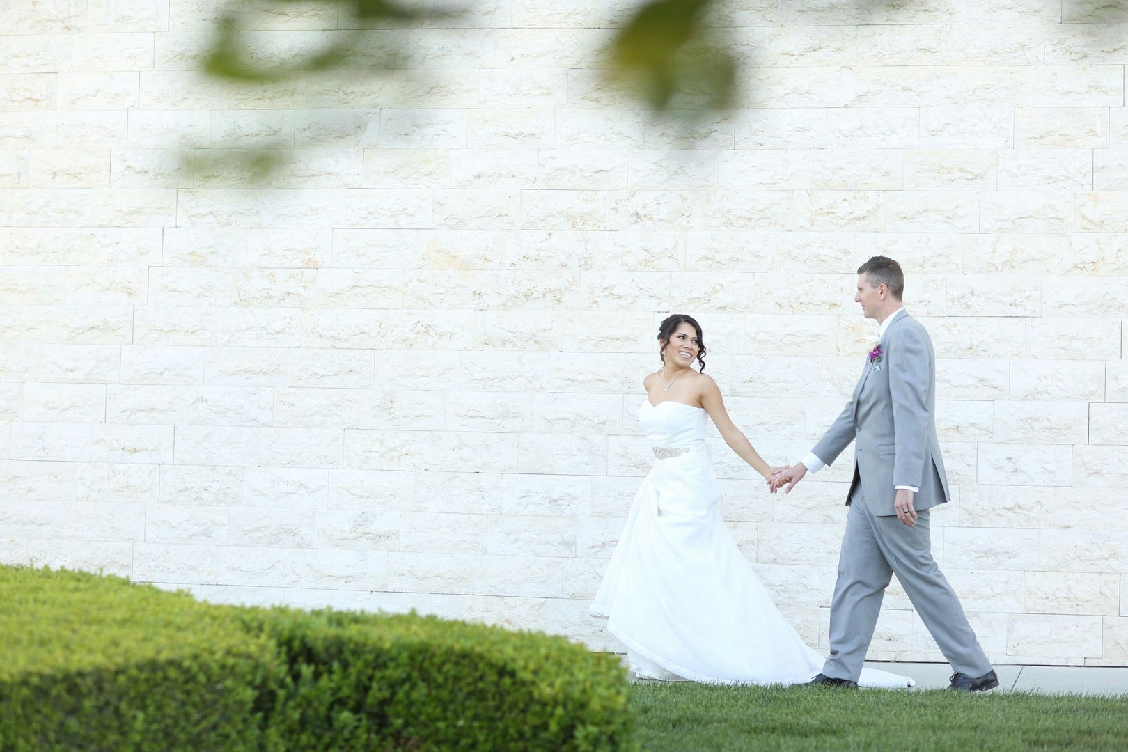 A bride and groom are walking together, holding hands, in front of a light-colored stone wall.