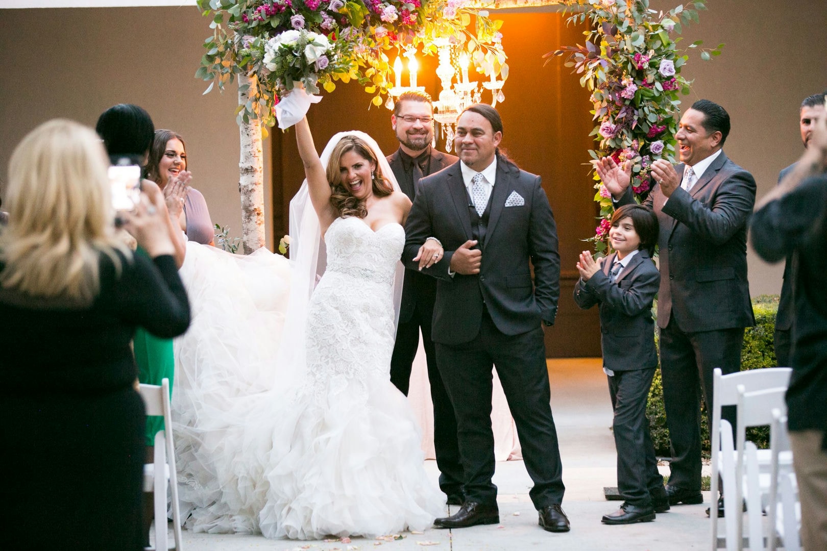 A wedding ceremony with the bride and groom, guests in suits, and a floral archway.