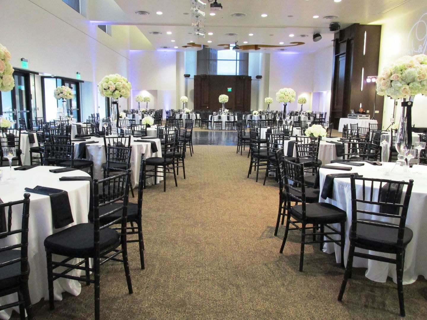 Large room with many black chairs and round tables. White tablecloths, floral centerpieces.