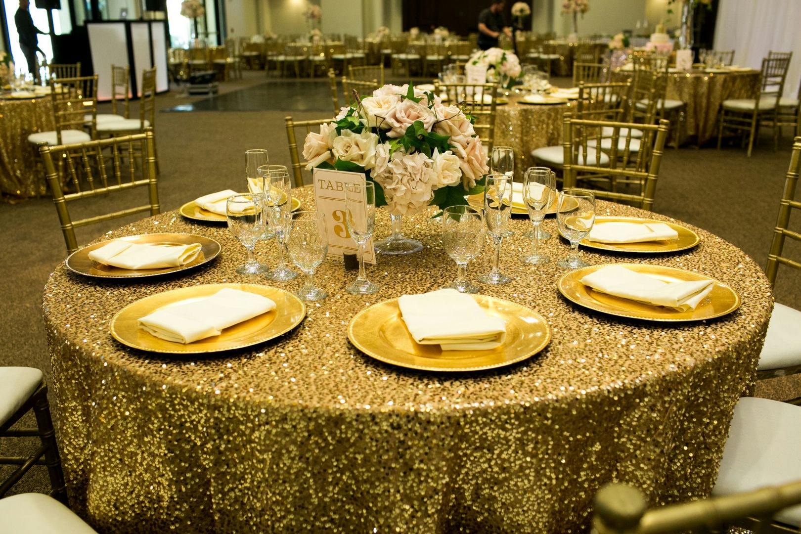 Gold sequin tablecloths, round gold plates, and light-colored napkins on tables with chairs. Floral centerpiec