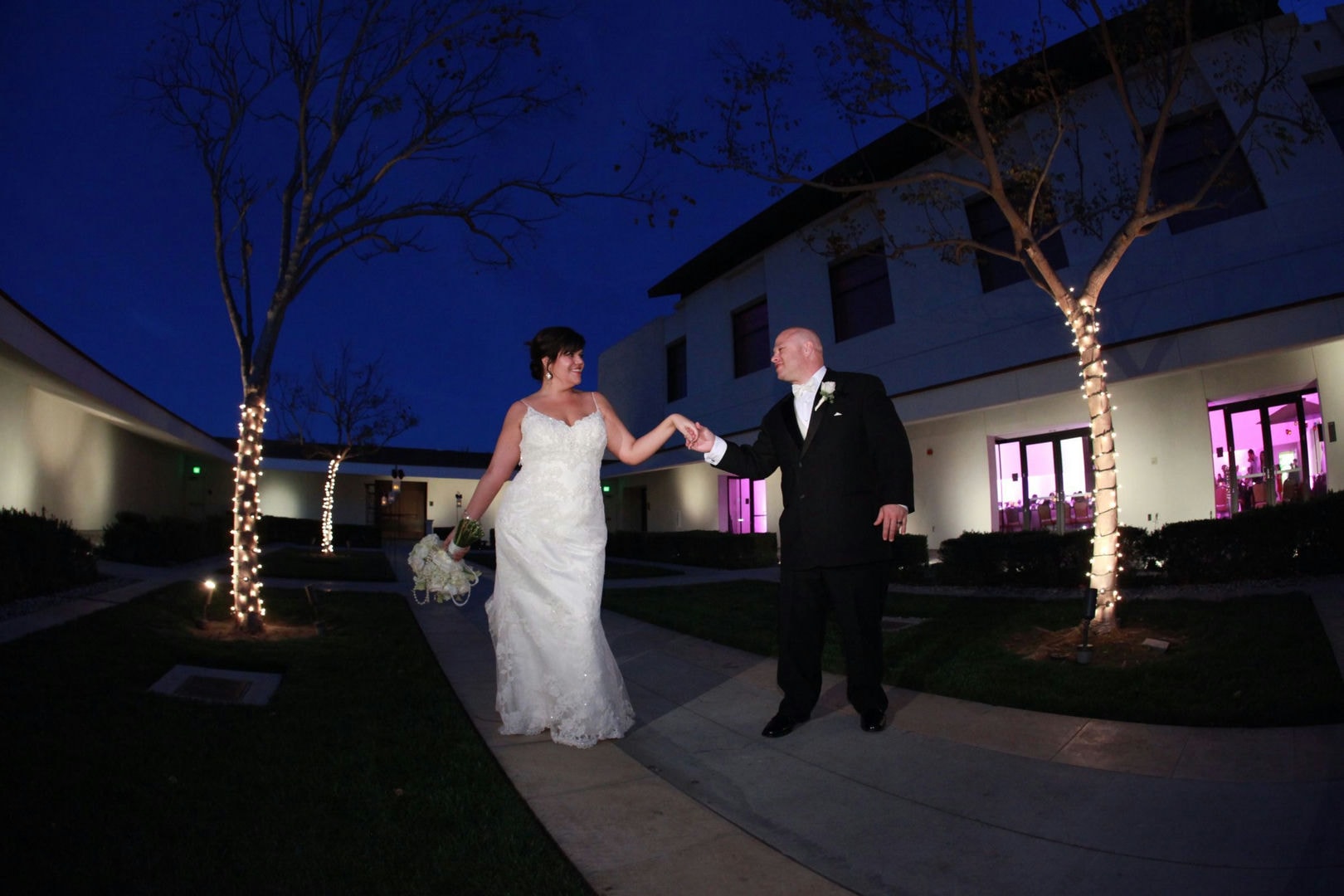 A bride and groom are walking hand-in-hand outside at night. The scene is lit by string lights.