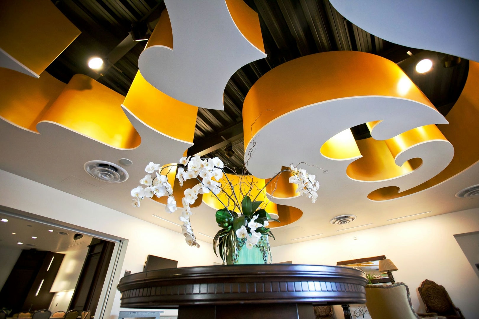 A room with a decorative, curved, yellow and gray ceiling, a round wooden table, and a vase of white orc