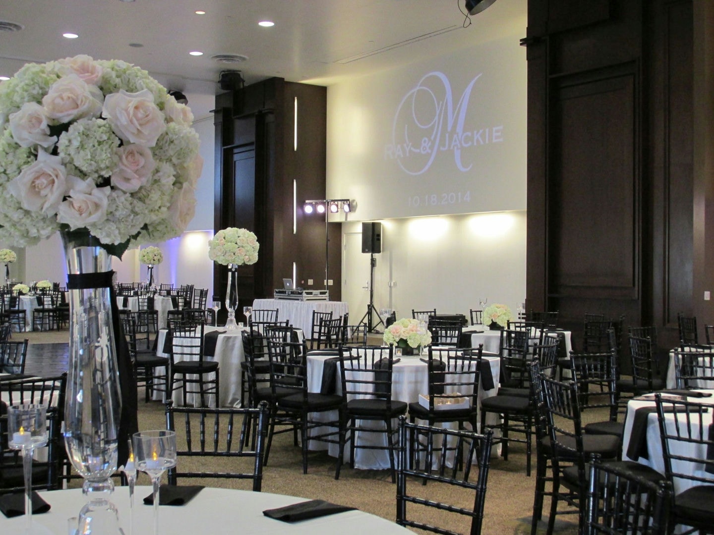 A banquet hall with round tables, black chairs, and white linens. Floral centerpieces are visible.