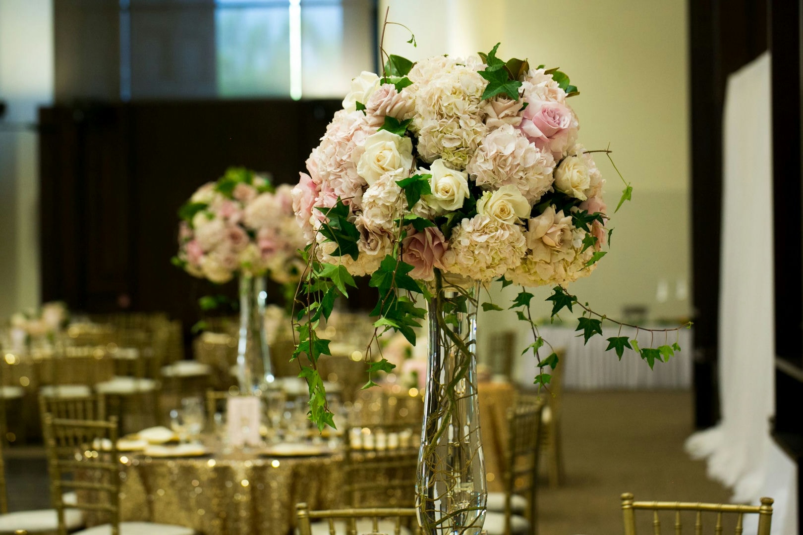 A banquet hall with tables, chairs, and tall centerpieces of flowers.