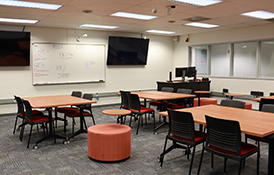 A bright computer classroom with white tables, blue chairs, and monitors. Windows and a chalkboard are visible.